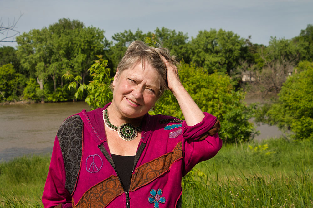 Photo: Di Brandt in red shirt running her hand through her hair in front of a river and treed background.